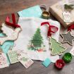Christmas-themed flat lay with gift tags, painted wooden ornaments, a white cloth bag with a Christmas tree print, a wrapped present with a bow, green and red paint, and holiday cards on a wooden surface.
