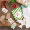 Handmade Christmas gift tags, festive cards, and paint jars on a wooden table. Tags feature words like “JOY,” “BELIEVE,” and “HOHOHO.” Wrapped gift and Santa-themed item are also visible.