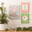 Three festive holiday signs with stripes and Christmas designs hang on a white wall above a wooden table decorated with a potted evergreen and ornaments.