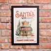 Framed sign on brick wall reads “Santa’s Workshop” with a festive house, Christmas tree, and gifts. Text below says “Toys and Candy Since 1823” and “Candy Cane Lane.”.