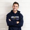 Smiling person with short brown hair wearing a navy blue “BELIEVE IN BELIEVE Ted Lasso” hoodie, standing with arms crossed in front of a light wooden wall.