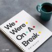 A spiral notebook with “We Were On A Break” in bold letters and colored dots, referencing the TV show Friends, lies next to a teal mug filled with coffee on a gray surface.
