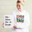 A smiling woman in a white hoodie holds a notebook that says, “We Were On A Break.” Her hoodie features a colorful “How you doin’?” graphic, referencing the TV show Friends.