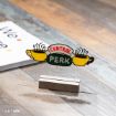 A small acrylic sign with the “Central Perk” logo from the TV show Friends, featuring three steaming yellow coffee cups, stands on a wooden base atop a rustic table.