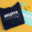 A navy hoodie with “BELIEVE IN BELIEVE” and “TED LASSO” printed on it is folded on a yellow surface, next to a blue paper with the same text, a paint container, and a round blue pin.