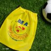 A yellow drawstring bag with “BE A GOLDFISH” and a goldfish illustration sits on green grass next to a black and white soccer ball.