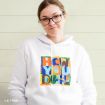 Woman with glasses wearing a white hoodie featuring colorful text that says “How You Doin?” with a cartoon frame design, standing in front of a light wooden wall.