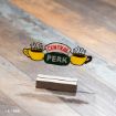 A clear sign with the “Central Perk” logo, featuring three steaming yellow coffee cups, stands on a wooden base atop a light wood surface.