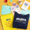 Ted Lasso-themed merchandise arranged on a yellow background, including a navy hoodie, yellow drawstring bag, posters, stickers, a mug, and paint pots, all featuring motivational phrases and goldfish graphics.