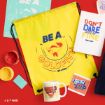 Flat lay of Ted Lasso-themed items on a red surface, including a yellow drawstring bag with “Be a Goldfish,” a mug, stickers, a notebook, and paint containers.