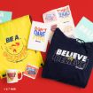 Ted Lasso-themed merchandise displayed on a red surface, including a yellow drawstring bag, a navy sweatshirt, a mug, a notebook, and several colorful stickers with motivational quotes.