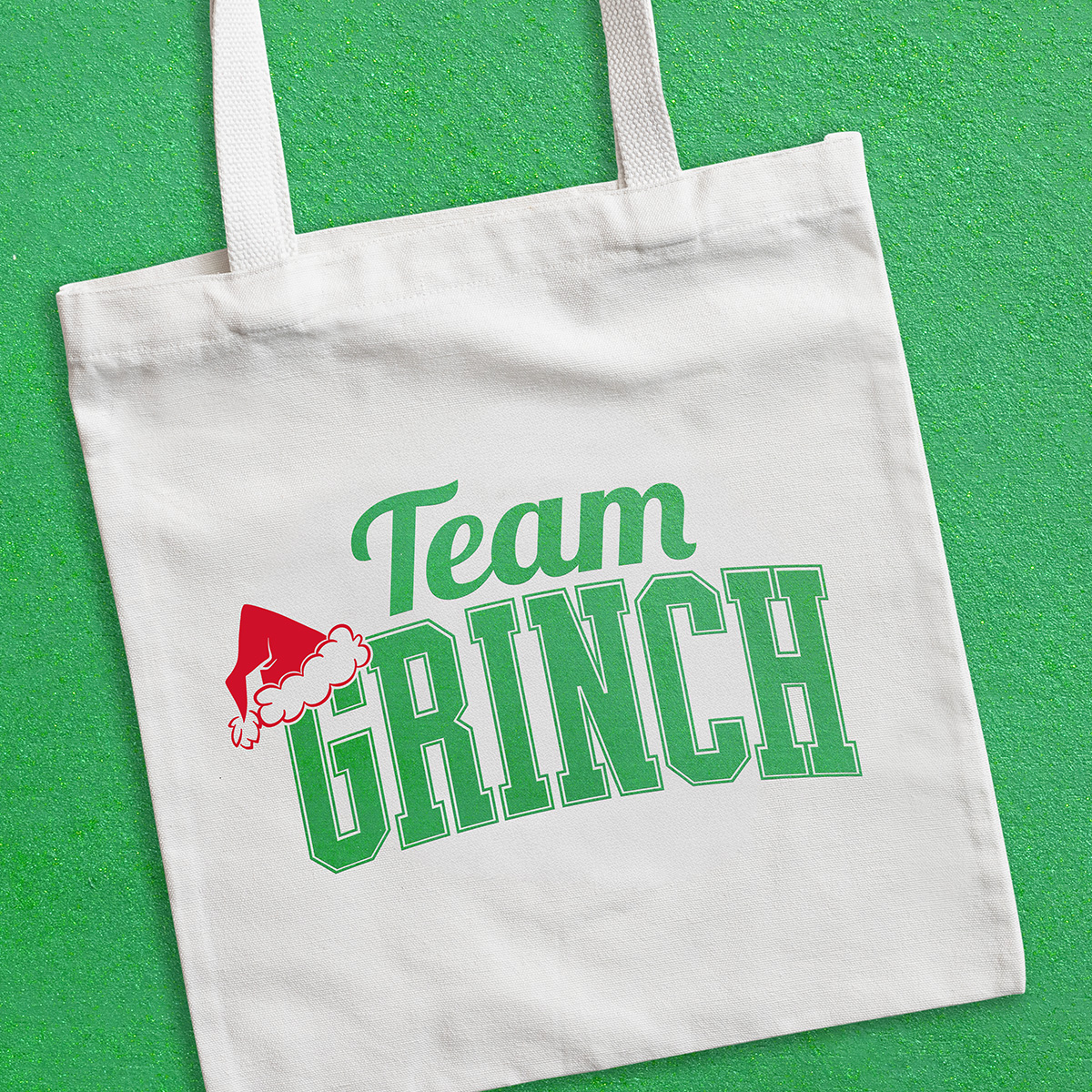 A white tote bag on a green surface with “Team GRINCH” in green letters, featuring a red Santa hat on the “G.”.