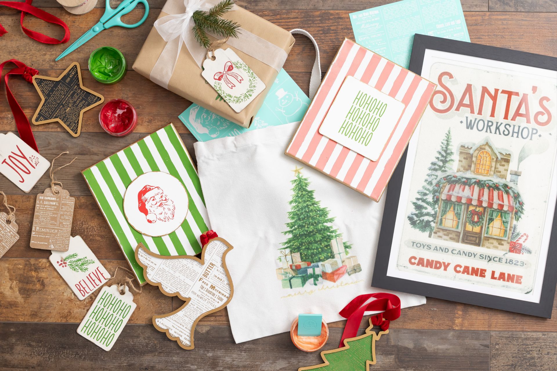 A collection of Christmas-themed craft supplies, including a wrapped gift, a white tote bag with a tree print, and a framed “Santa’s Workshop” sign on a wooden surface.