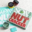 A wooden craft sign reading “Son of a Nutcracker” with a matching turquoise stencil, two jars of colored chalk paste, and a small scraper.