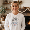 A woman wearing a white sweatshirt featuring an illustration of a snowman wearing a top hat and the text “Snow much fun!”.