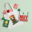 A white tote bag printed with Buddy the Elf surrounded by two wooden signs reading “Raised by Elves” and “Son of a Nutcracker,” accompanied by small gift boxes and ornaments.