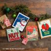 Holiday decor on a wooden floor including a framed Frosty the Snowman print, a National Lampoon’s Christmas Vacation sign, a A Christmas Story leg lamp print, an Elf “Son of a Nutcracker” sign, and two wrapped gifts among pine branches.