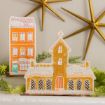 Two decorative wooden gingerbread-style house ornaments, one tall and orange and one shorter church building, dusted with artificial snow and surrounded by greenery.