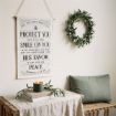 A hanging canvas sign printed with the Bible verse Numbers 6:24-26, displayed on a wall above a wooden console table with a lit candle, greenery, and a green pillow.