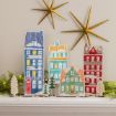 A holiday display on a mantel features four illustrated cardboard buildings and three small trees decorated with faux snow, with two gold star ornaments on the wall above.