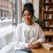 A person wearing a sweatshirt with a candle design sits at a cafe table holding a mug of coffee next to a book.