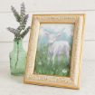 A framed painting of a white lamb in a field of flowers, sitting next to a green glass vase with sprigs of lavender.
