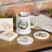 White mug with a hedgehog illustration holding two cinnamon sticks, surrounded by a fox block, a squirrel block, an owl coaster, and a deer coaster on a wooden table.