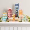 A collection of six colorful, flat wooden tabletop house decorations with faux-snow accents, arranged on a white mantelpiece with green garland.