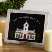 Framed sign depicting a church with snow on its roof, reading “MAY THE LORD BLESS YOU” surrounded by two lit pillar candles.