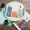 A circular wooden tray featuring three illustrated houses, with a matching stencil, pots of paint, a scraper, and a tea towel printed with the same house designs.