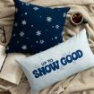 A blue square pillow with white snowflakes, a light blue rectangular pillow with the text “UP TO SNOW GOOD,” an open book, and a mug of tea resting on a textured blanket.
