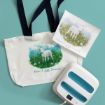A white canvas tote bag and a paper print, both featuring a watercolor illustration of a lamb in a field with the text “Now I Am Found Luke 15:6-7,” next to a white and blue heat press machine.