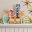 A collection of colorful, illustrated wooden house and church cutouts with faux snow decoration, displayed on a white mantelpiece with gold star ornaments on the wall above.