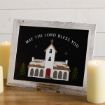 A framed sign featuring an illustration of a snow-covered church and the text “MAY THE LORD BLESS YOU,” displayed between two lit pillar candles.