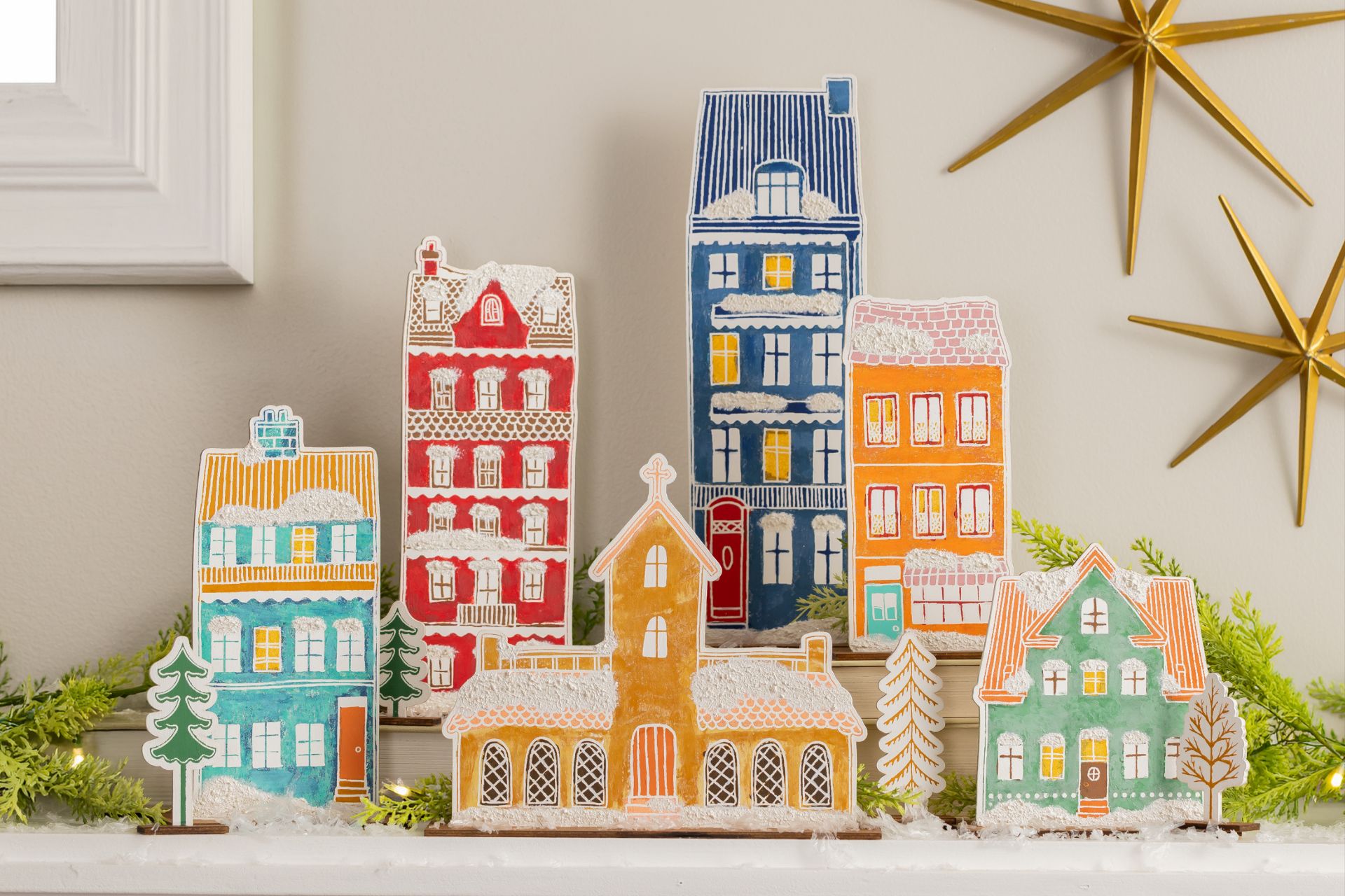 A collection of flat, painted wooden cutout houses and a church arranged on a mantel, decorated with faux snow and gold star ornaments on the wall behind.